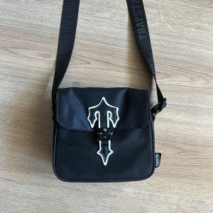 Svart axelväska från Trapstar - Säljer en äkta svart axelväska från Trapstar med justerbar rem och coolt spänne framtill. Väskan har meshdetaljer på framsidan och tydlig Trapstar-logga på både rem och etikett. Perfekt storlek för det viktigaste och riktigt street i stilen. Köpt på Trapstars hemsida.