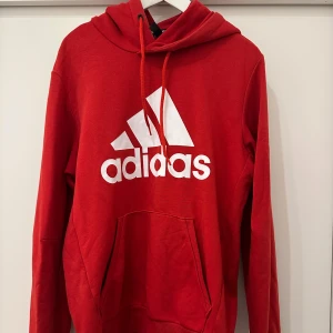 Röd Adidas hoodie med logga - Riktigt snygg röd hoodie från Adidas med stor vit logga framtill. Klassisk känguruficka, huva med snörning och långa ärmar. Perfekt för dig som gillar sportig och avslappnad stil. Mjuk och skön i bomullsmix.