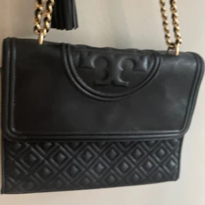 Svart quiltad axelväska från Tory Burch - Snygg svart axelväska från Tory Burch i skinn med quiltad nederdel och kedjerem i guld. Väskan har en dekorativ tofs och det klassiska Tory Burch-loggan framtill. Perfekt storlek för det viktigaste och ger en lyxig känsla till din outfit.