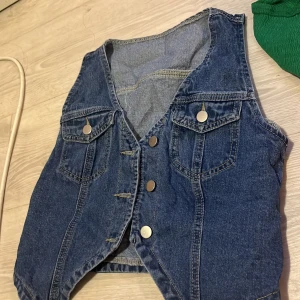 Klassisk jeansväst med knappar - Snygg blå jeansväst med fyra silvriga knappar framtill och två bröstfickor med lock. Västen har en v-ringad skärning och markerade sömmar som ger en cool vintagekänsla. 