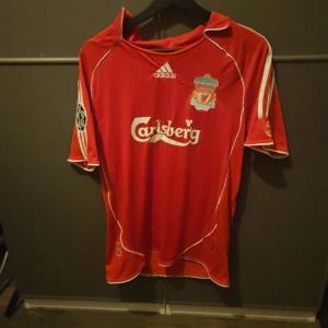 Liverpool Crouch 15 Adidas fotbollströja - Röd Liverpool fotbollströja med vita detaljer från Adidas. Tryck med Carlsberg på bröstet, klubbmärke och Champions League-märke på ärmen. Nummer 15 och Crouch på ryggen. Klassisk passform med korta ärmar och vita ränder på axlarna. Perfekt för fans av Liverpool och retrotröjor.