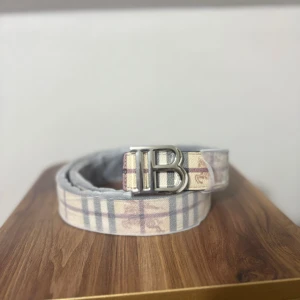 Rutmönstrat Burberry-inspirerat bälte - Snyggt bälte med klassiskt rutmönster i beige, grått och vinrött samt en stor silvrig B-spänne framtill. Bältet har en sida i svart med diskret rutmönster och insida i beige rutigt. Perfekt accessoar för att lyfta din outfit.