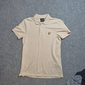 Beige pikétröja från Lyle & Scott - Snygg beige pikétröja från Lyle & Scott med klassisk krage och tre knappar framtill. Tröjan har det ikoniska gula örnloggan broderad på bröstet och är gjord i mjuk bomull. Perfekt för en clean och stilren look.