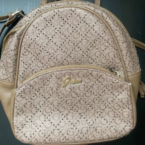 Beige ryggsäck från Guess med logomönster - Inköpspris 1800 Snygg beige ryggsäck från Guess med klassiskt logomönster och guldfärgade detaljer. Väskan har flera fack, dragkedjor och är tillverkad i syntetmaterial med skinnliknande känsla. Perfekt storlek för vardagens alla prylar och har både handtag och justerbara axelremmar.