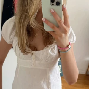 Vit blus med puffärm och knappar - Supersöt vit blus med puffiga korta ärmar och söt volangkant nertill. Blusen har en djup rundad urringning och dekorativa knappar framtill. Perfekt för dig som gillar romantisk och trendig stil. Tillverkad i mjuk bomull som känns skön mot huden.