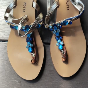 Guldiga sandaler med blå stenar Motta - Snygga sandaler från Motta i guldigt skinn med remmar och dekorativa blå stenar framtill. Handgjorda i Italien med platt sula och öppen tå. Perfekta för att ge din sommarlook extra bling och färg.