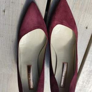 Vinröda mockapumps från Rupert Sanderson - Snygga pumps i vinröd mocka från Rupert Sanderson. Skorna har spetsig tå och elegant klack, perfekta för att lyfta din outfit. Lyxig känsla med mjukt material och klassisk design.