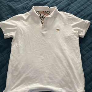 Vit pikétröja från Burberry - Snygg vit pikétröja från Burberry med klassiskt rutigt tyg på insidan av kragen och knäppningen. Tröjan har korta ärmar, broderad logga på bröstet och är gjord i 100% bomull. Perfekt för dig som gillar stilrena och tidlösa plagg. Passar både S och M