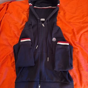 Mörkblå Moncler hoodie med dragkedja - Snygg mörkblå hoodie från Moncler med hel dragkedja, huva och vita/röda detaljer på ärmar och huva. Klassisk logga på armen. Perfekt för dig som gillar streetwear och exklusiva märken.