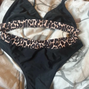 Svart bikini med leoparddetaljer, M - Snygg svart bikini i storlek M med breda leopardmönstrade band både upptill och nedtill. Bikinin har en enkel, sportig form med trekantsöverdel och höga trosor. Materialet är mjukt och stretchigt, perfekt för stranden eller poolen.