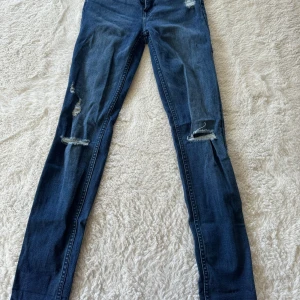 Mörkblå skinny jeans från H&M - Snygga mörkblå skinny jeans från H&M med slitna detaljer på benen. Klassisk femficksmodell med dragkedja och knapp framtill. Perfekta för en cool och avslappnad stil.