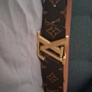 Louis Vuitton monogram skärp brun/guld - Säljer ett Louis Vuitton skärp i klassiskt monogrammönster med brun bas och guldiga LV-detaljer. Bältet har en ikonisk LV-spänne i metall och är gjort i exklusivt skinn. Perfekt accessoar för att lyfta din outfit med lyxig känsla. Använda 2 gånger