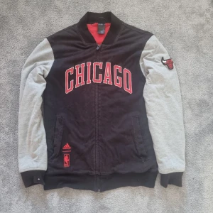 Chicago Bulls bomberjacka från Adidas - Snygg svart och grå bomberjacka från Adidas med Chicago Bulls-tryck och broderad logga på ärmen. Jackan har dragkedja, röda detaljer och quiltat rött foder. Perfekt för dig som älskar basket och streetstyle.