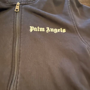 Svart hoodiejacka från Palm Angels - Svart hoodiejacka från Palm Angels med dragkedja och stor logga i vitt både fram och bak. Jackan har huva med metallöljetter och kängurufickor. Tillverkad i mjuk bomull, perfekt för en chill streetwear-look.
