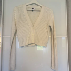 Vit ribbad kofta från H&M Divided - Superfin vit ribbad kofta från H&M Divided i storlek S. Koftan är croppad med v-ringning och har tre knappar framtill. Långärmad och stickad i mjukt material, perfekt att slänga över en topp eller klänning.