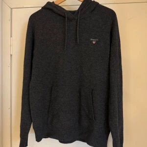 Mörkgrå cashmere hoodie från GANT - Mörkgrå hoodie från GANT med broderad logga på bröstet. Klassisk modell med huva och snörning samt känguruficka framtill. Mjuk och skön, perfekt för chill dagar. Passar dig som gillar stilren och enkel design.