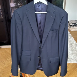 Mörkblå kostym från These Glory Days || Nypris 3000kr  - Snygg mörkblå kostym från These Glory Days med klassisk kavaj och matchande byxor. Kavajen har två knappar, tre fickor fram och slät baksida. Kostymen är tillverkad i ull och har ett stilrent, tidlöst snitt som passar perfekt till mer uppklädda tillfällen.