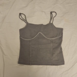 Croppat linne - Säljer ett grått croppat linne med tunna justerbara axelband och markerad söm över bysten. Säljer då det inte kommer till användning. Skriv om ni har några frågor!