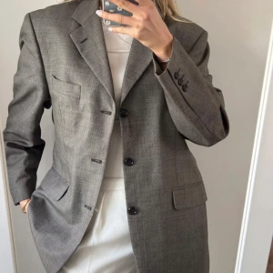 Grå kavaj i ullmix - Stilren grå kavaj med diskret rutigt mönster och klassisk passform. Kavajen har tre knappar framtill, fickor med lock och en bröstficka. Perfekt för en dressad look och passar snyggt till både jeans och kostymbyxor.