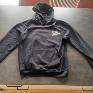 Svart hoodie från Warp med tryck - Svart hoodie från Warp med vit text 'WATER SNOW CONCRETE' på bröstet. Klassisk känguruficka framtill och huva. Mjuk insida och ribbade muddar vid ärmslut och nederkant. Perfekt för chill dagar eller när du vill ha en enkel streetstyle.