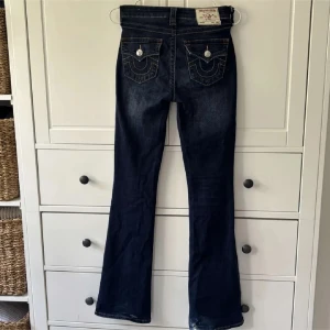 True Religion mörkblå bootcut jeans - Mörkblå jeans från True Religion med bootcut passform och klassiska kontrastsömmar. Snygga fickdetaljer bak med stora silverfärgade knappar och tydlig logopatch i midjan. Jeansen är i denim med en något utsvängd benvidd och cool vintagekänsla. Se slitage på sista bilderna. Storlek 25. Modell Becca 