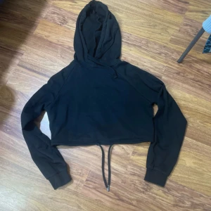 Svart croppad hoodie från NA-KD - Svart croppad hoodie från NA-KD med långa ärmar och huva. Tröjan har snörning i nederkanten för justerbar passform och är tillverkad i mjuk bomullsmix. Perfekt för en chill och avslappnad stil.
