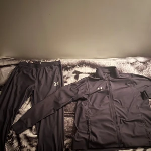 Svart träningsset från Under Armour - Säljer ett svart träningsset från Under Armour med byxa och jacka. Setet har en stilren look med dragkedja på jackan och logga på både jacka och byxa. Materialet är mjukt och stretchigt, perfekt för träning eller chill. Byxorna har resår i midjan för skön passform.