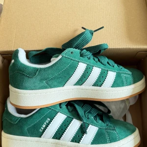 Adidas Campus gröna sneakers mocka - Adidas Campus sneakers i grön mocka med klassiska vita ränder och vit sula. Skorna har snörning och en platt gummisula i ljusbrun färg. Ikonisk retrostil med rund tå och extra gröna samt vita skosnören medföljer.