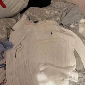 Vit kabelstickad tröja Polo Ralph Lauren - Vit kabelstickad tröja från Polo Ralph Lauren med klassisk blå logga broderad på bröstet. Tröjan har rund halsringning och långa ärmar, perfekt för lager-på-lager. Snygg och tidlös design som funkar året runt.