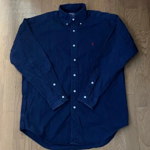 Ralph lauren skjorta  -  Strlk S-M Klassisk mörkblå skjorta från Polo Ralph Lauren i 100% bomull. Skjortan har långa ärmar, knappar framtill och ikonisk röd broderad logga på bröstet. Perfekt för dig som gillar stilrena och tidlösa plagg med snygga detaljer.