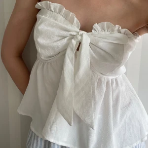 Vit axelbandslös topp med rosett - Supersöt vit axelbandslös topp med volangkant upptill och stor knytrosett framtill. Toppen har bandeau-modell och smockad rygg för extra komfort. Lätt och luftig, perfekt för varma dagar. 🥰🥰