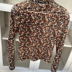Blommig mesh polotopp  - Säljer en tunn och stretchig mesh polotopp från New Yorker i storlek S. Toppen har ett blommigt mönster i orange, brunt, vitt och gult på svart botten. Långärmad och figurnära passform, perfekt att styla lager på lager eller ha som en elegant blus/ topp