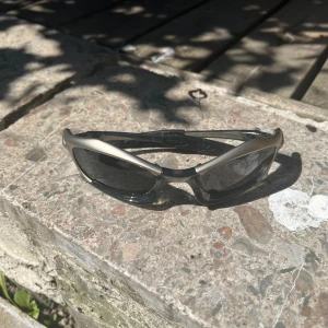 Oakley X-Metal solglasögon silver - Säljer ett par Oakley X-Metal solglasögon med mattsilverfärgad båge och svarta glas. Sportig och futuristisk design med markerade linjer och ikonisk Oakley-logga på sidan. Perfekta för dig som vill sticka ut med en cool och unik look.