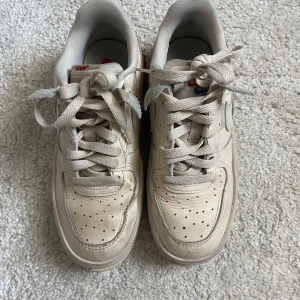 Nike Air Force 1 Shadow beige sneakers - Nike Air Force 1 Shadow i beige med röda detaljer på sulan och svarta sömmar bak. Klassisk siluett med snörning, perforerad tå och dubbla lager på swoosh och häl. Skorna är i skinn och har en chunky platt sula för extra attityd.