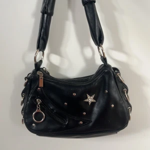 Svart axelväska med stjärndetalj - Cool svart axelväska i skinnimitation med silvriga metalldetaljer, dragkedjor och en glittrig stjärna framtill. Väskan har bred axelrem, ringar på sidorna och flera fack med dragkedja. Perfekt för dig som gillar edgy och trendig stil.