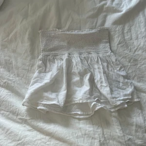 Vit bandeau tubtopp i linne - Supersöt vit bandeau tubtopp med smock upptill och volangig nederdel. Gjord i lätt och luftigt linnematerial, perfekt för varma dagar. Enkel att matcha med jeans eller kjol för en chill sommarlook. Från Gina Young i 158/164