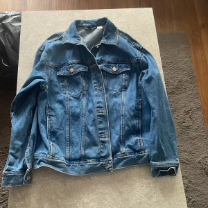 Klassisk blå jeansjacka - Snygg blå jeansjacka med klassisk passform och silverfärgade knappar. Jackan har två bröstfickor med lock och knappar samt två sidofickor. Perfekt att slänga över en hoodie eller t-shirt för en chill look.