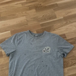 Grå Moncler t-shirt med patch - Snygg grå t-shirt från Moncler med rund hals. På bröstet sitter en stor broderad Moncler-patch i vitt, svart och rött. T-shirten är i mjuk bomull och har en clean look. Prickarna är vatten droppar mitt hår är blött. Priset kan diskuteras