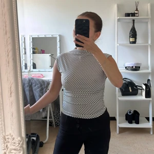 Randig svartvit ärmlös topp - Säljer en slim, ärmlös topp med svartvita diagonala ränder. Toppen har hög hals och rynkade detaljer längs sidorna för en snygg passform.
