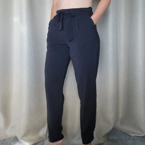 Svarta kostymbyxor med knyt i midjan - Snygga svarta kostymbyxor med hög midja och knytband. Byxorna har raka ben, sidfickor och resår baktill för extra komfort. Perfekta för en clean och stilren look.