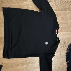 Svart Moncler sweatshirt med logga - Svart sweatshirt från Moncler med klassisk rund hals och broderad logga på bröstet. Tröjan har långa ärmar och en snygg detalj med röd, vit och blå rand på insidan av kragen. Perfekt för en clean och stilren look.