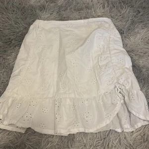 Vit broderad kjol från H&M - Superfin vit kortkjol från H&M med broderade blommönster och volangkant. Kjolen har snörning på ena sidan och är gjord i mjuk bomull, perfekt för sommarens alla äventyr. Elastisk midja för skön passform.