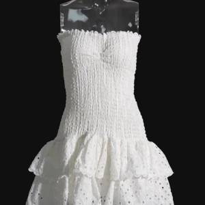 Vit axelbandslös kortklänning med volang - Supersöt vit kortklänning med smockad överdel och axelbandslös design. Kjolen har två lager volanger med broderade hål och spetsdetaljer. Perfekt för sommarens alla tillfällen, ger en fräsch och najs sommar vibe. Köpte för 850kr, pris kan diskuteras 💗