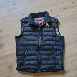 Svart dunväst från Parajumpers - Snygg svart dunväst från Parajumpers med quiltad design och dragkedja framtill. Västen har två fickor med dragkedja och en hög krage med gul detalj. Superlätt och perfekt för lager-på-lager-stil. Insidan är ljusgrå och materialet är syntet.