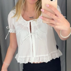 Vit blus - Supersöt vit blus med korta ärmar, spetsdetaljer vid krage och nederkant samt knyt framtill. Blusen har volangkant och broderade detaljer som ger en romantisk vibe. Perfekt för dig som gillar en feminin och trendig look.