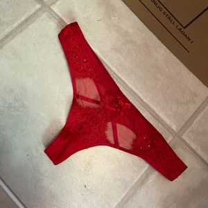 Röda spetsstring med rosett och strass strl M - Snygga röda stringtrosor i mesh och spets med blommigt mönster, liten rosett framtill och glittrande strassdetaljer. Trosorna har tunna band och transparenta partier för en extra stylish look. Obs lappen är bortklippt 