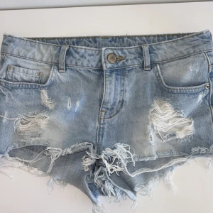 Ljusblå slitna jeansshorts - Säljer ett par ljusblå jeansshorts med slitna detaljer och fransig kant. Shortsen har klassisk femficksmodell, låg midja och är gjorda i denim. Perfekta för en avslappnad och trendig look i sommar. 