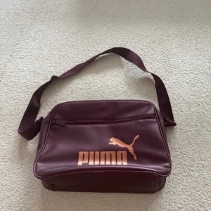 Lila träningsväska - Säljer en  lila träningsväska från Puma med justerbar rem och stort logotryck i brons färg på framsidan. Väskan har dragkedja, ett stort huvudfack och extra fack framtill. 