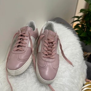 Valentino Garavani rosa sneakers - Snygga sneakers från Valentino Garavani i ljusrosa och vitt skinn. Klassisk modell med snörning, rund tå och coola nitar på sulan baktill. Perfekta för dig som vill ha en stilren men ändå unik look. Mjuka rosa skosnören och kontrasterande vit sula.