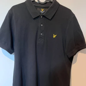 Svart pikétröja från Lyle & Scott - Klassisk svart pikétröja från Lyle & Scott med gul broderad logga på bröstet. Tröjan har krage, tre knappar framtill och är kortärmad. Tillverkad i bomull för en skön och stilren look. Perfekt för dig som gillar enkel och snygg stil.